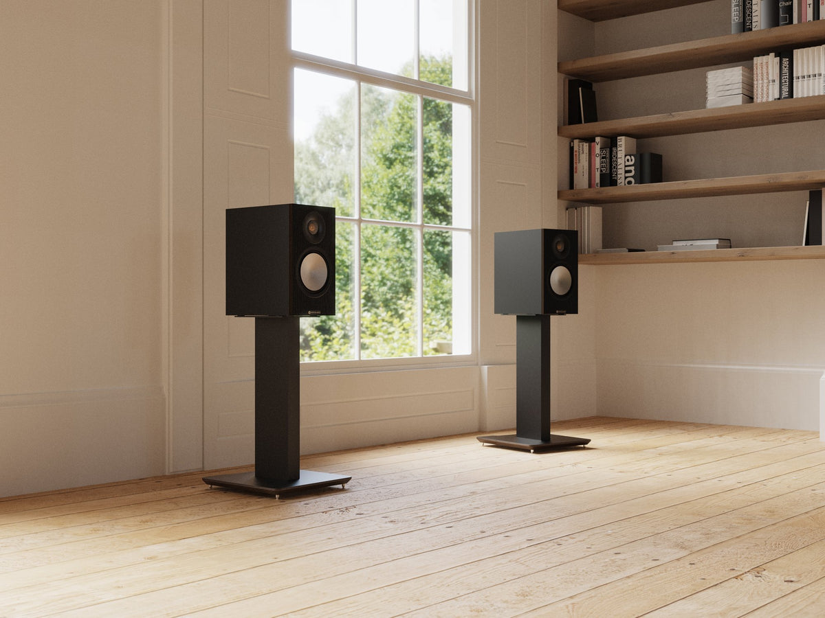 Two black floor-standing bookshelf speakers on slim black stands, placed in a bright room with light wood floors, a large window, and built-in wooden shelves.