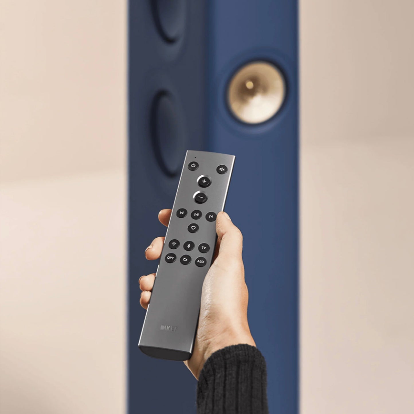 Hand holding a remote control in front of a blue speaker on a beige background