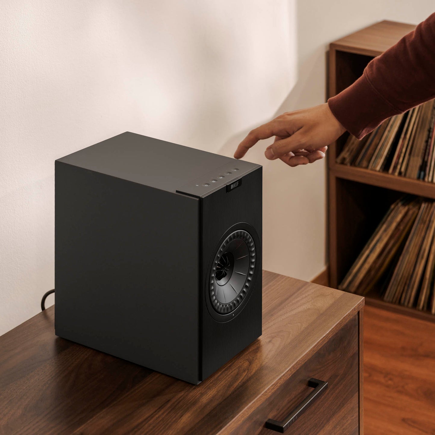 Black speaker on a wooden surface with a person reaching out to touch it, against a neutral background.