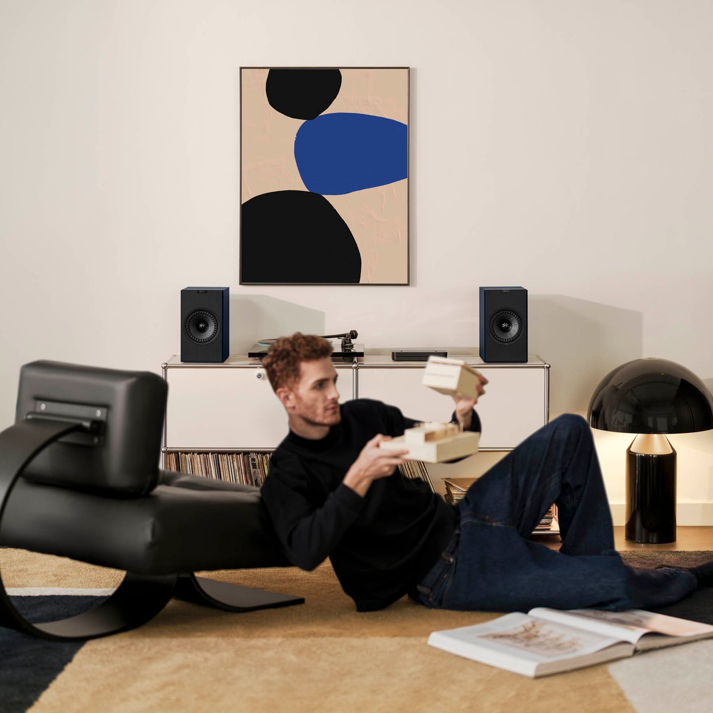 Man lying on a rug reading a book in a modern living room with abstract art and speakers.