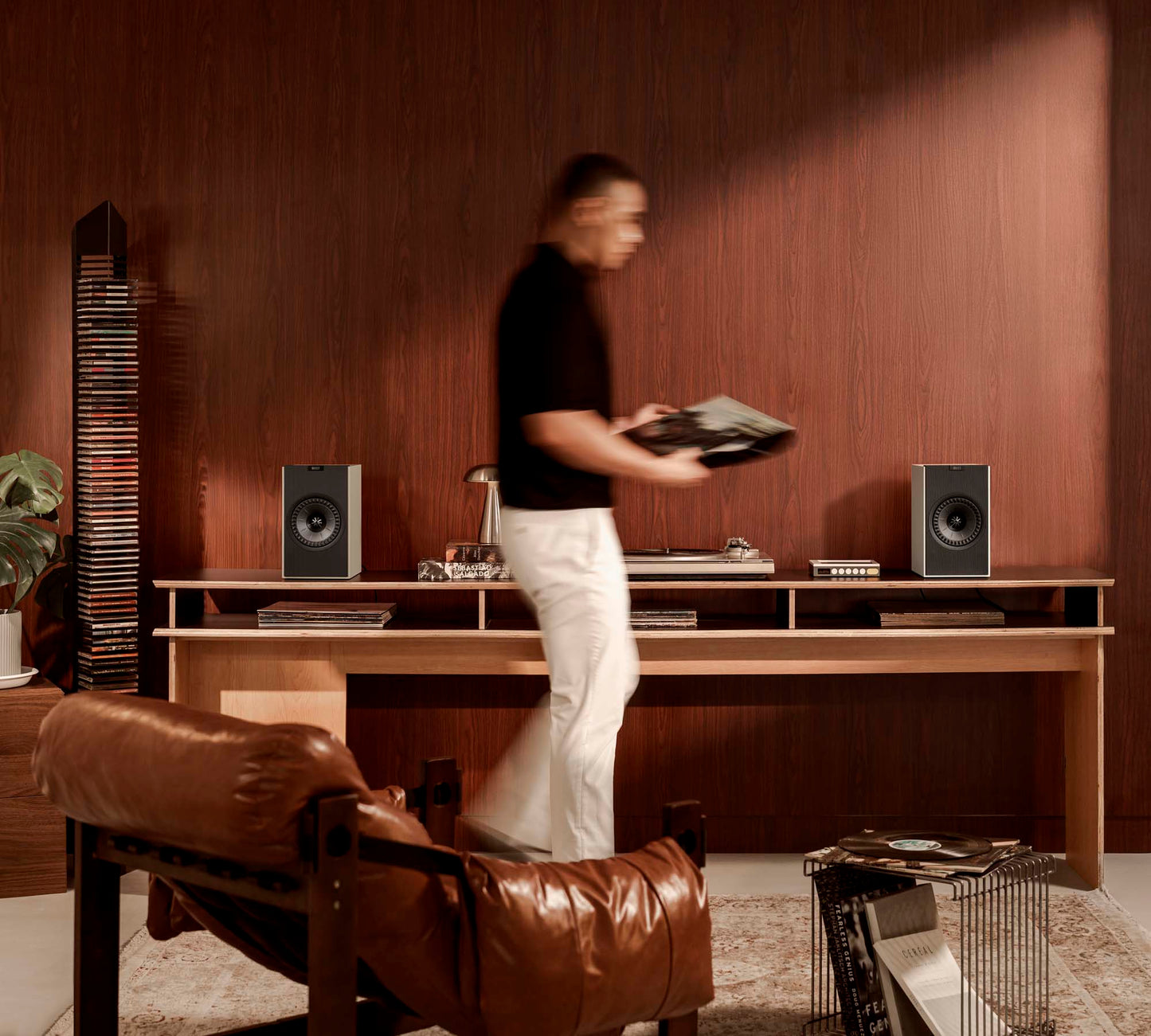 Person standing in a room with wooden walls, a brown leather chair, and a desk with electronic equipment.