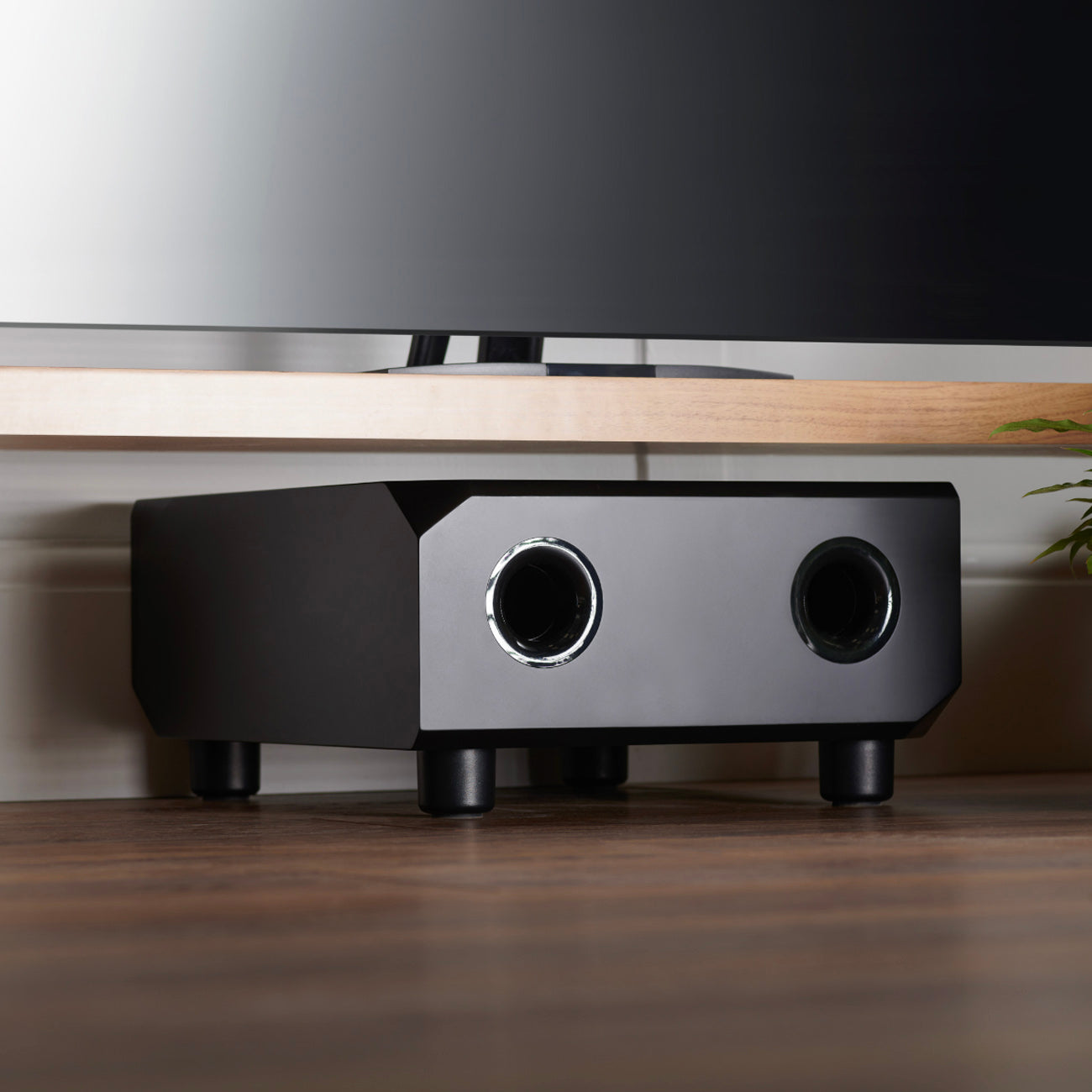 Black audio equipment under a wooden desk with a tv in the background