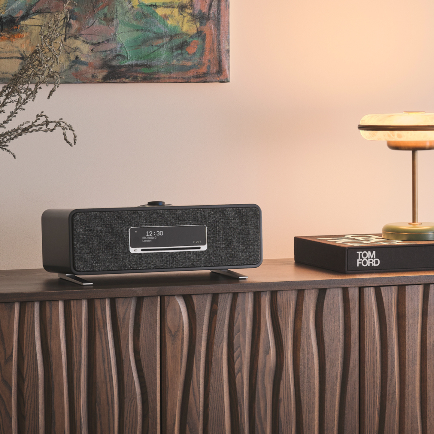 Modern clock radio on a wooden console table with a lamp and abstract painting in the background.