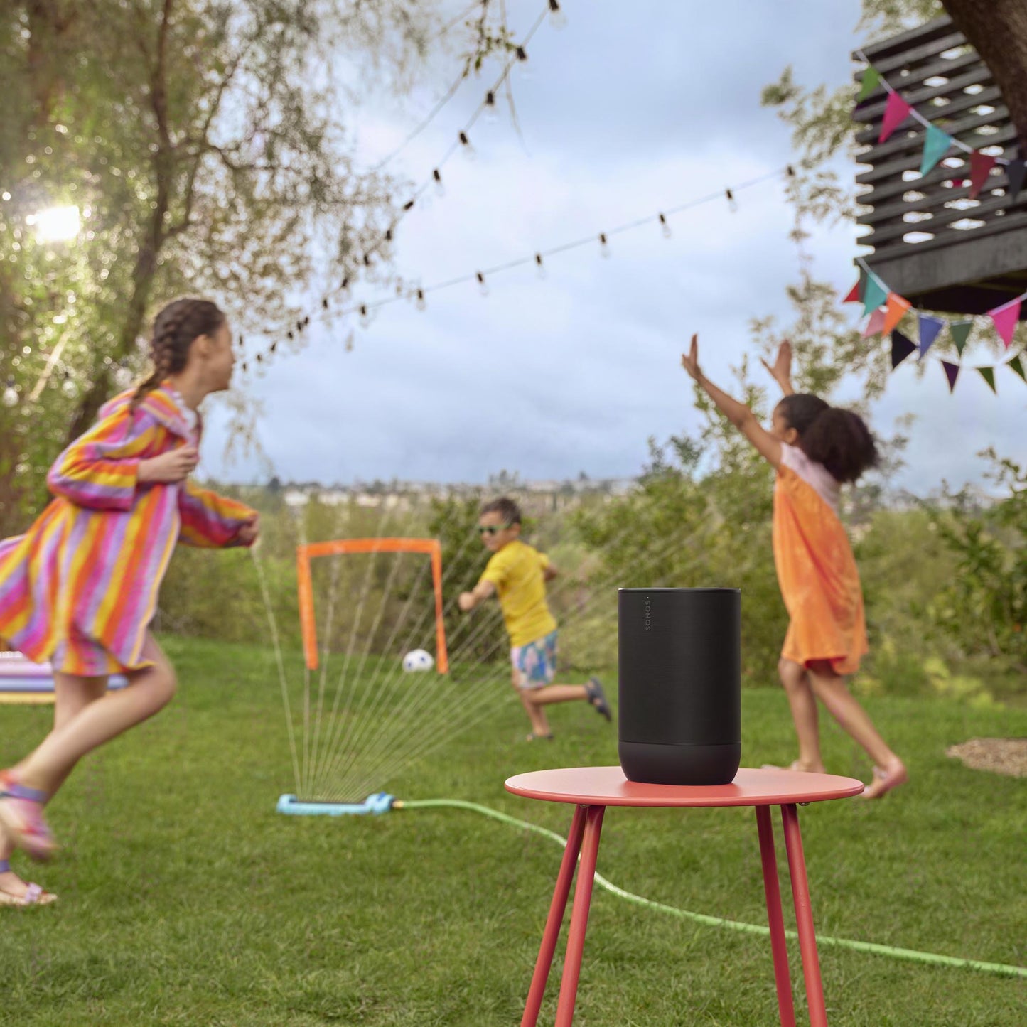 Black Sonos Move 2 on red table in garden with kids playing under sprinkler in backyard