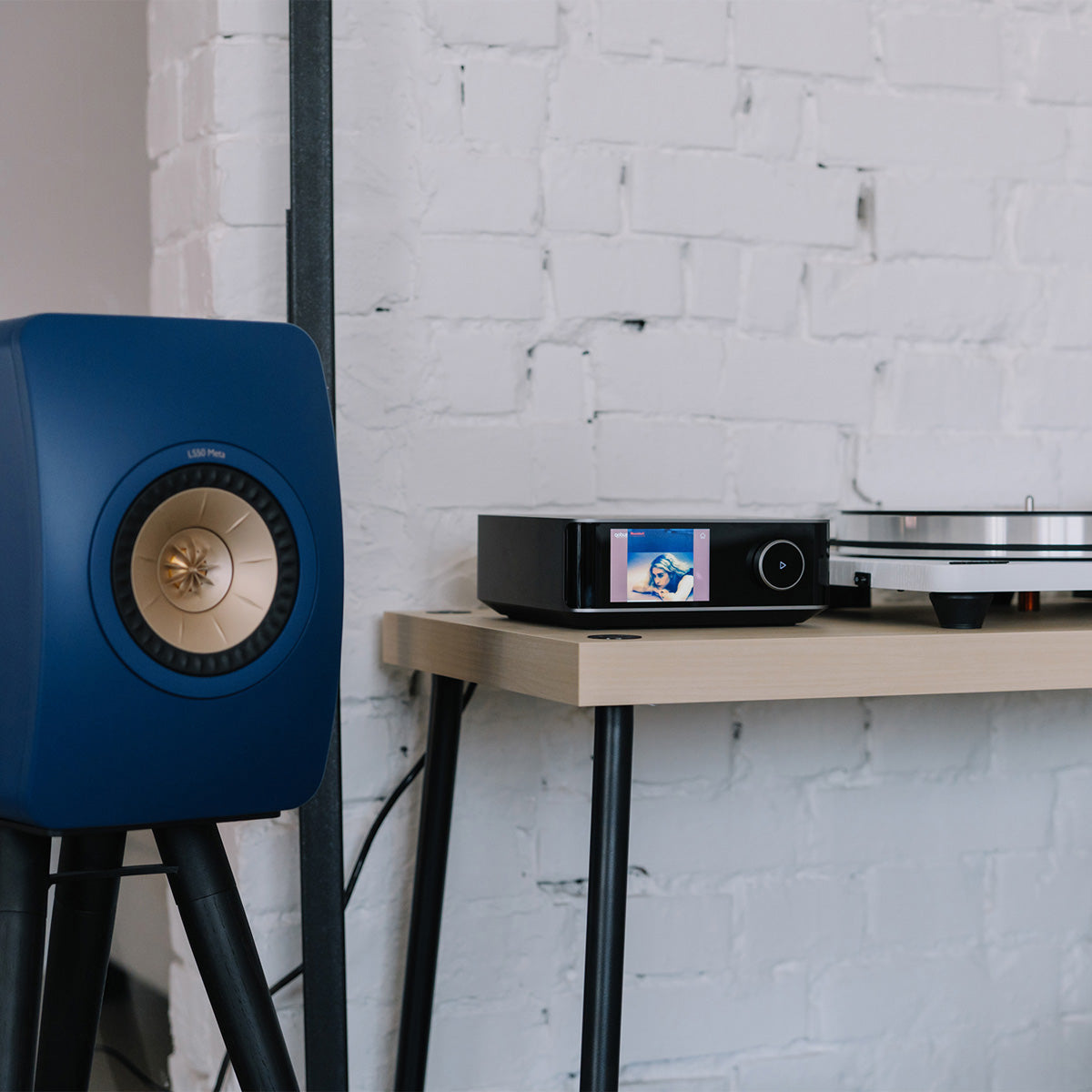 Blue speaker next to a turntable on a wooden table against a white brick wall.