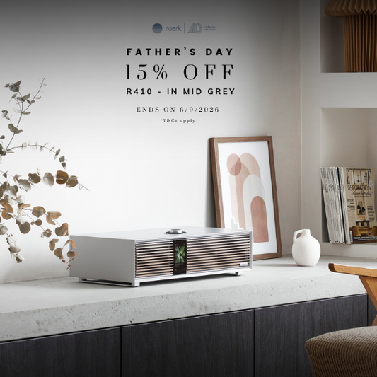 A silver speaker sits on a white marble console in a modern living room setting. The scene includes a terracotta vase with dried branches, a framed abstract print, books, a shelf with vases, and part of a brown armchair.