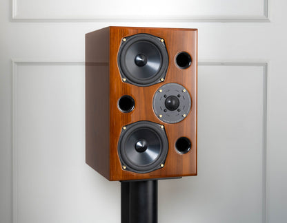 A wooden speaker cabinet with two large black woofers, a gray midrange driver, and small circular port holes, mounted on a black stand against a white paneled wall.