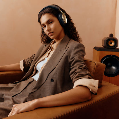 A woman with curly hair wearing over-ear headphones sits in a brown leather armchair, dressed in a brown blazer over a light-colored top. A wooden speaker is visible in the background against a warm beige wall.