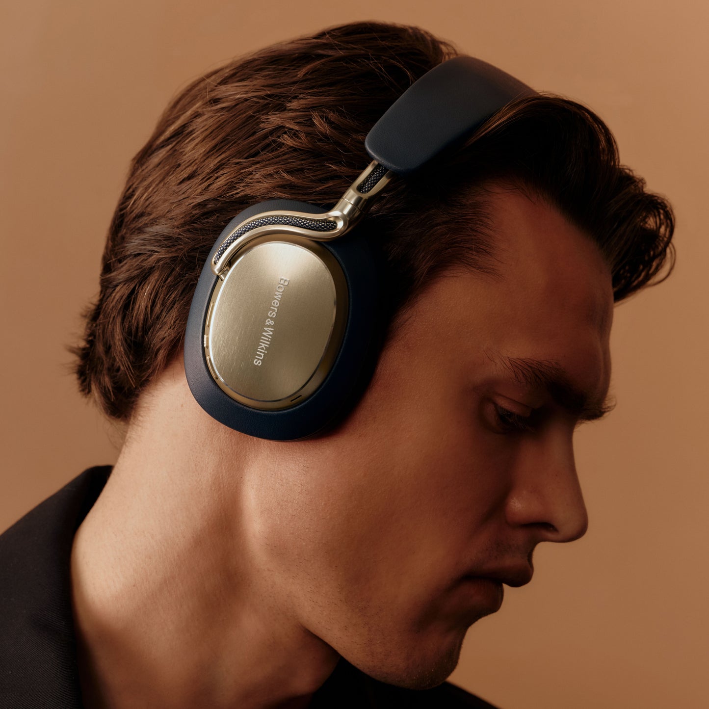 A close-up side profile of a man wearing over-ear headphones. The headphones have dark navy blue outer shells with metallic silver inner ear cups and a silver hinge