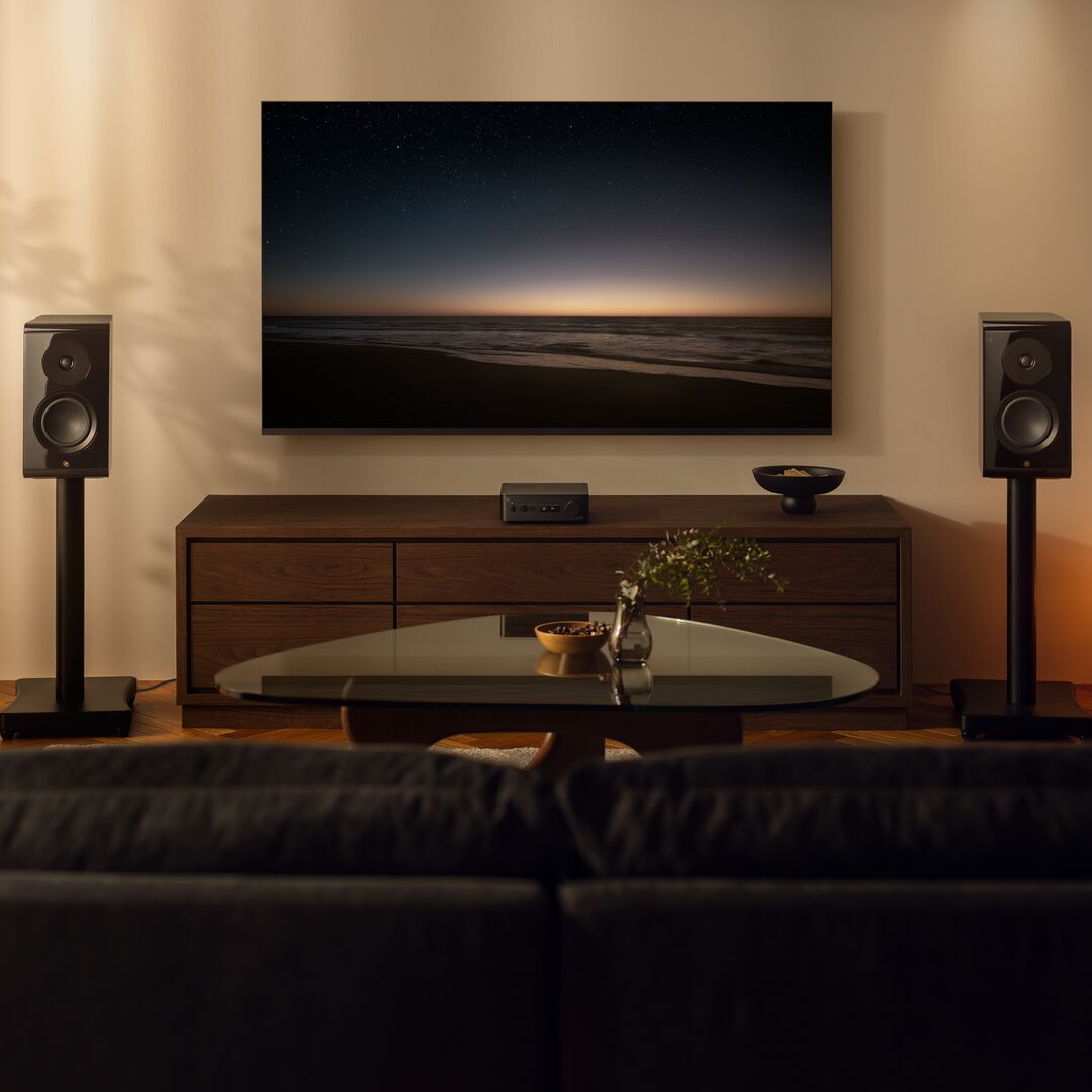 A large wall-mounted TV displaying a starry night sky over a beach, shown in a living room setup with a wooden media console, two black speakers on stands, a glass coffee table, and a dark sofa in the foreground.