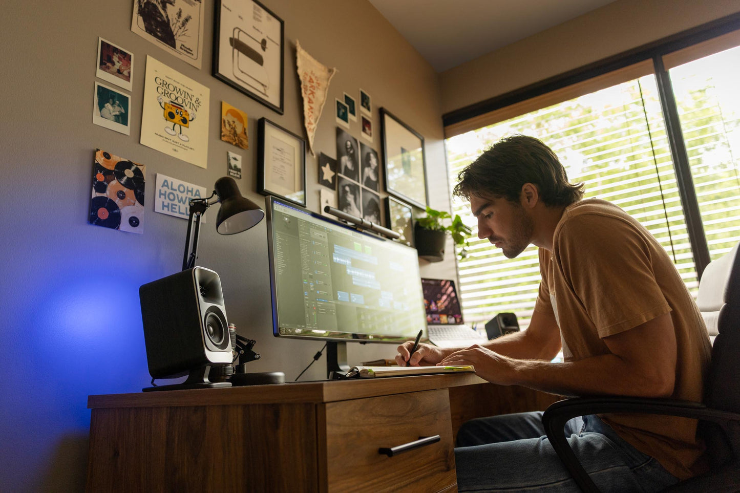 Person sitting at a desk with a computer and writing in a notebook, in a room with posters on the wall.