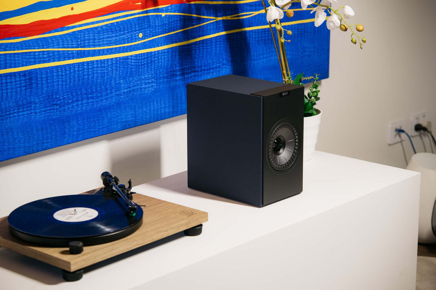 KEF Coda W Speaker and Audio Technica AT-LPW40WN Turntable on white shelf with bright artwork behind.