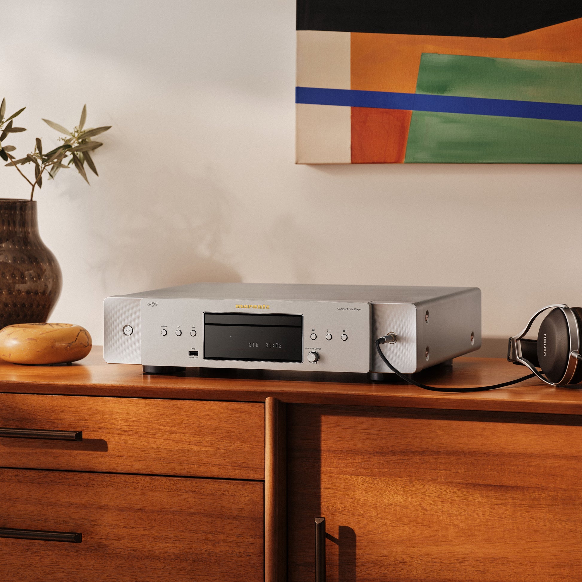 A silver Marantz CD 70 CD Player sits on a wooden cabinet in a styled home interior, with a vase of greenery,  and over-ear headphones nearby against a cream wall and abstract artwork.