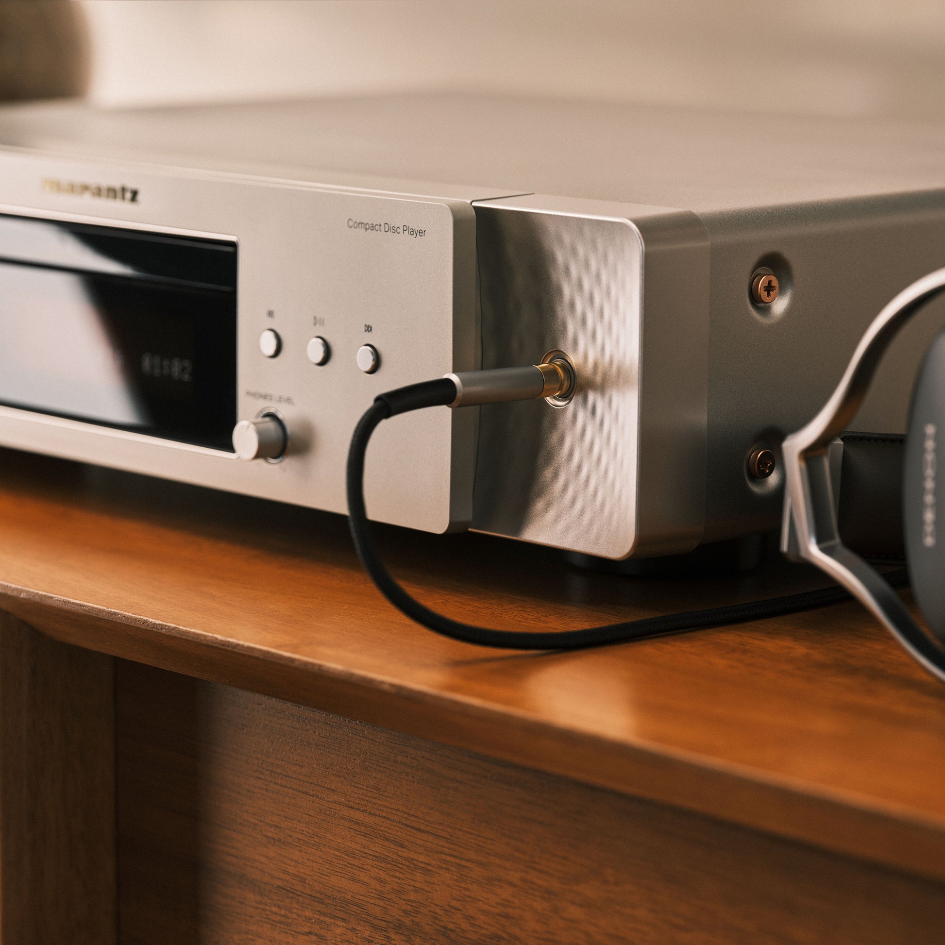 A close-up of a silver Marantz compact disc player sitting on a wooden shelf, with a black audio cable plugged in and part of a black and silver pair of headphones visible on the right.