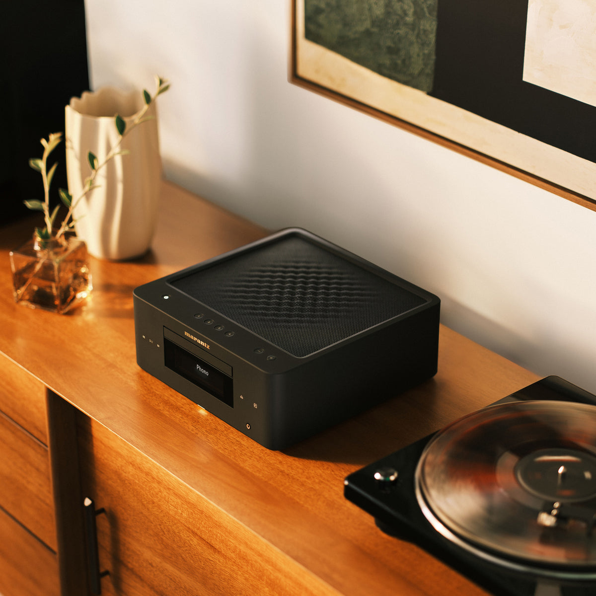 Marantz MCR-60N Compact All-In-One Hi-Fi System sits on a wooden cabinet in a styled interior setting, with a vase, framed wall art, and part of a turntable visible around it.