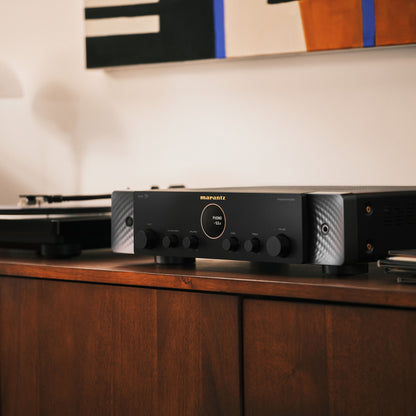 A black Marantz amplifier sitting on a dark wood media console in a home theater setting, with a turntable blurred in the background.