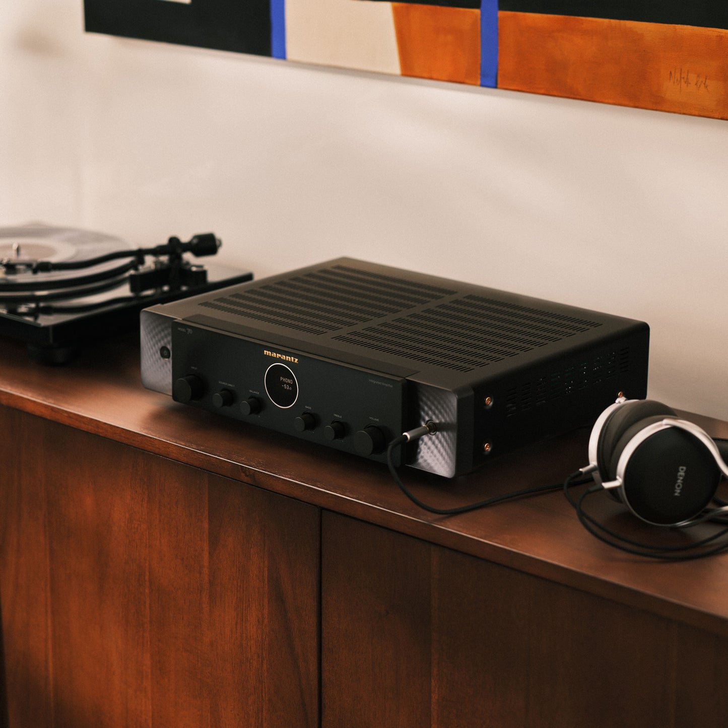 A black Marantz amplifier sits on a wooden cabinet in a home setting, with a turntable on the left and over-ear headphones on the right.