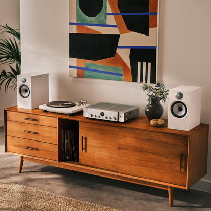 wooden media console in a warm brown finish, styled with a turntable, receiver, white speakers, and a vase against a beige wall with a colorful abstract painting.