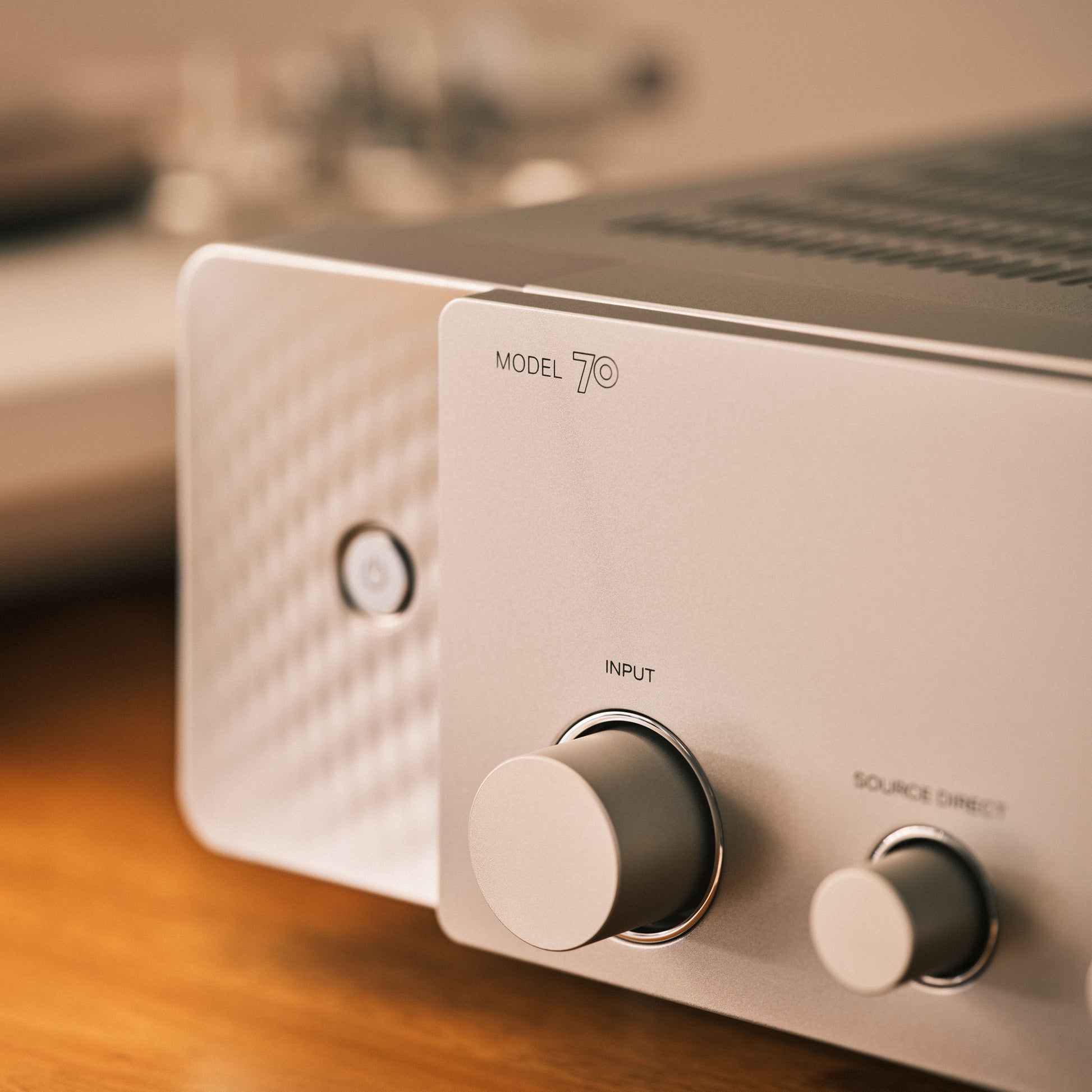 Close-up of a silver amplifier with front-panel controls, including a large rotary knob labeled INPUT and a smaller knob labeled SOURCE DIRECT, on a wooden surface.