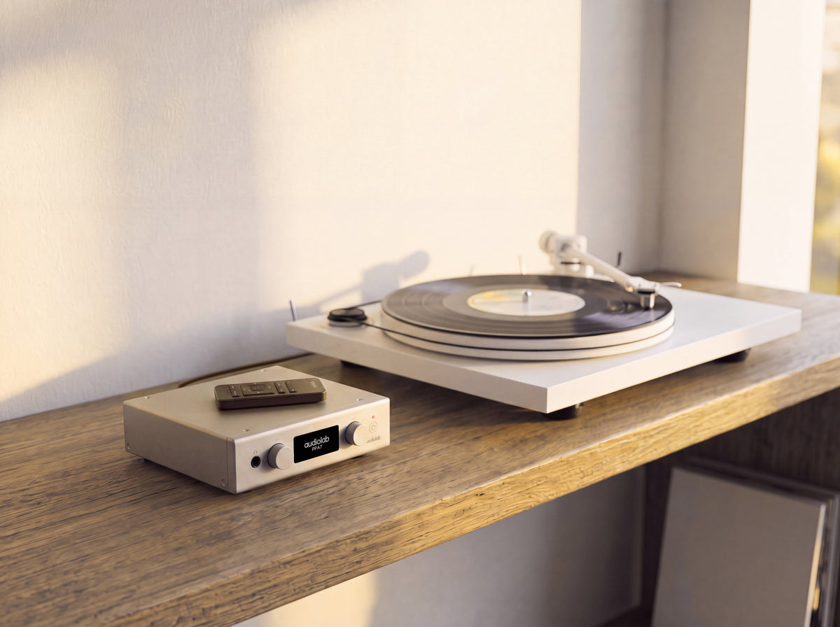 A modern turntable with a vinyl record on it sits on a wooden shelf next to a small silver audio receiver with a remote control, set against a warm, sunlit wall.