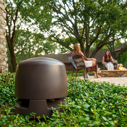 WiiM Outdoor Subwoofer on grass with people sitting by a fire pit in the background