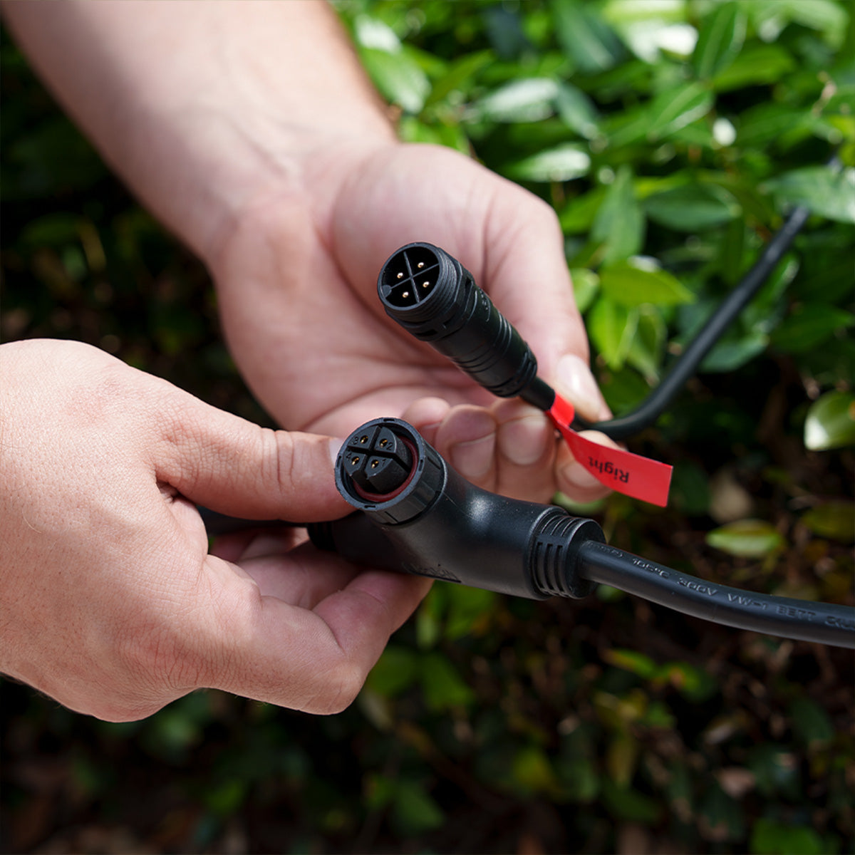 Hands holding two black connectors with red labels against a green leafy background