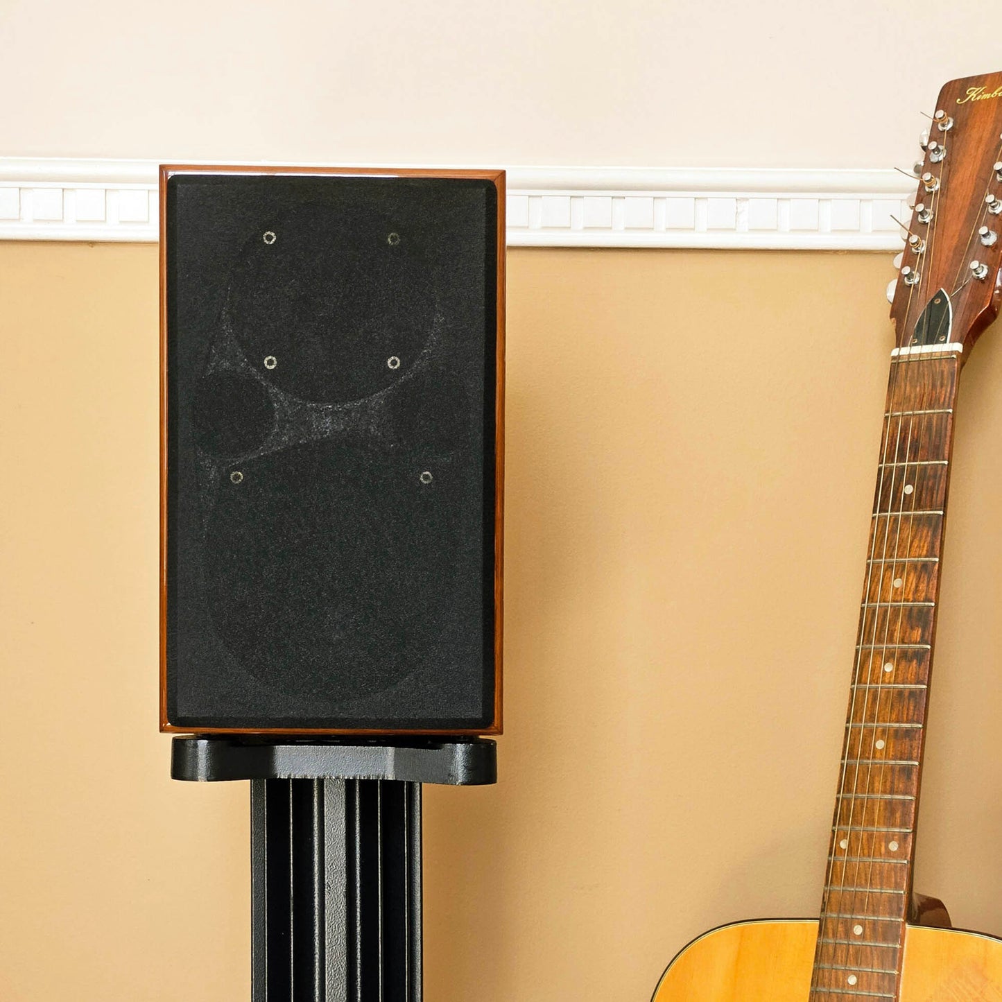 Acoustic Energy AE1 40th Anniversary Edition on a stand next to an acoustic guitar against a beige wall.