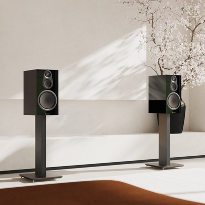 Two black bookshelf speakers on slim black stands, displayed in a minimalist modern room with soft sunlight and a flowering tree in the background.
