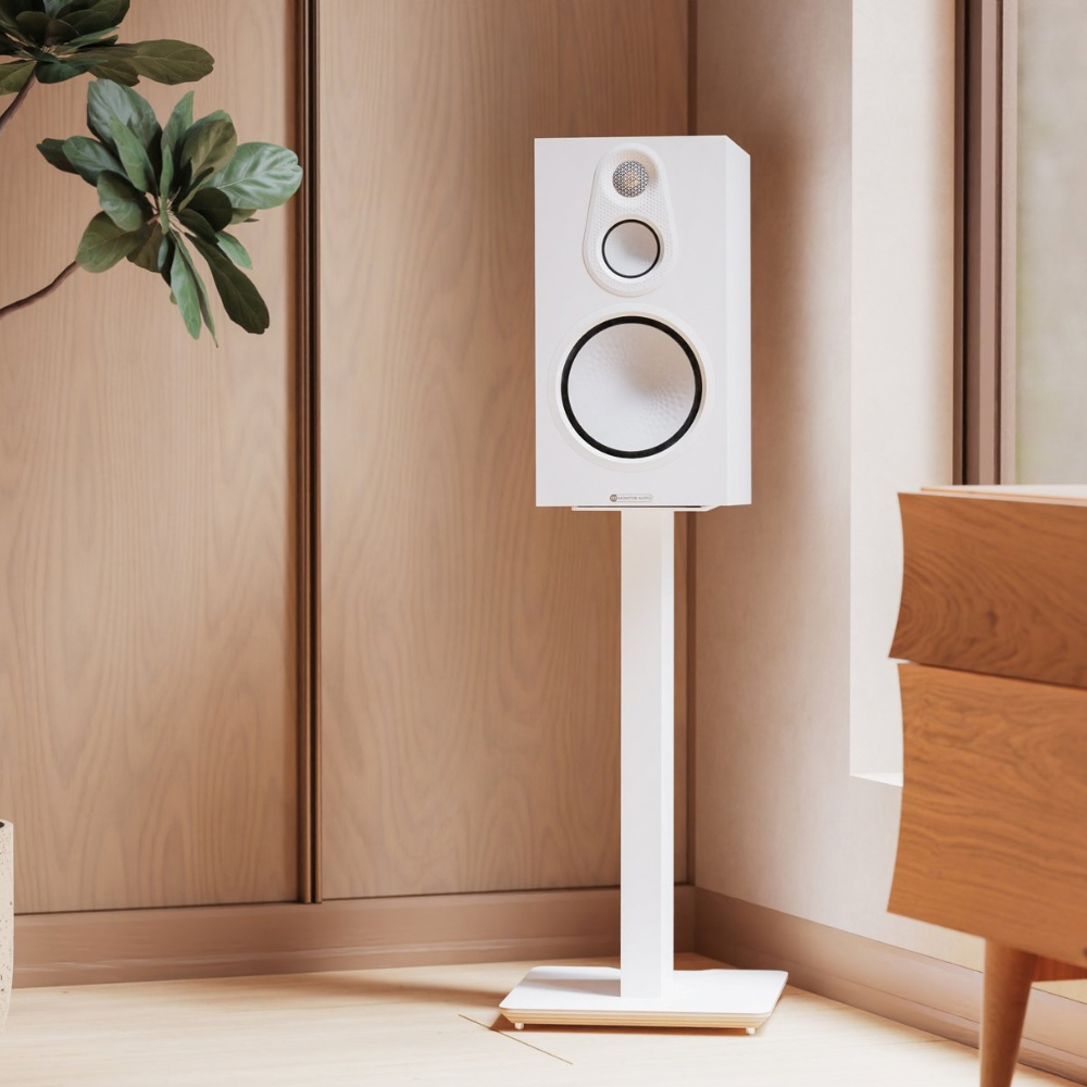 A white bookshelf speaker on a matching white stand, set in a minimalist room with light wood paneling and a wooden side table.