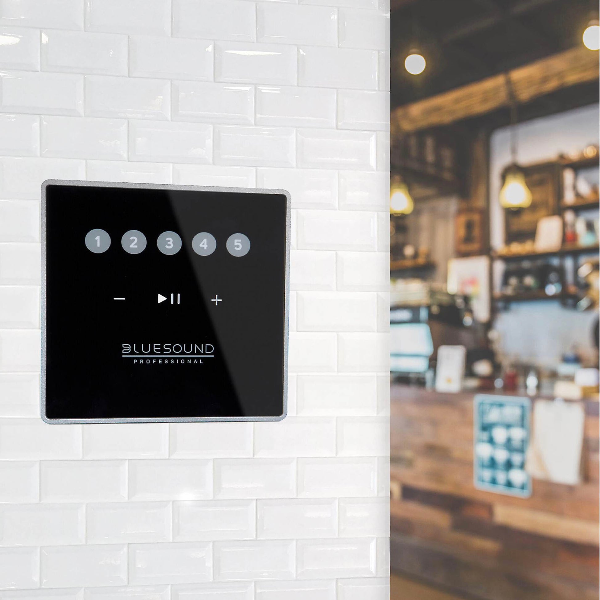 Black control panel with 'Bluesound' branding on a white tiled wall, blurred bar or café interior in the background.