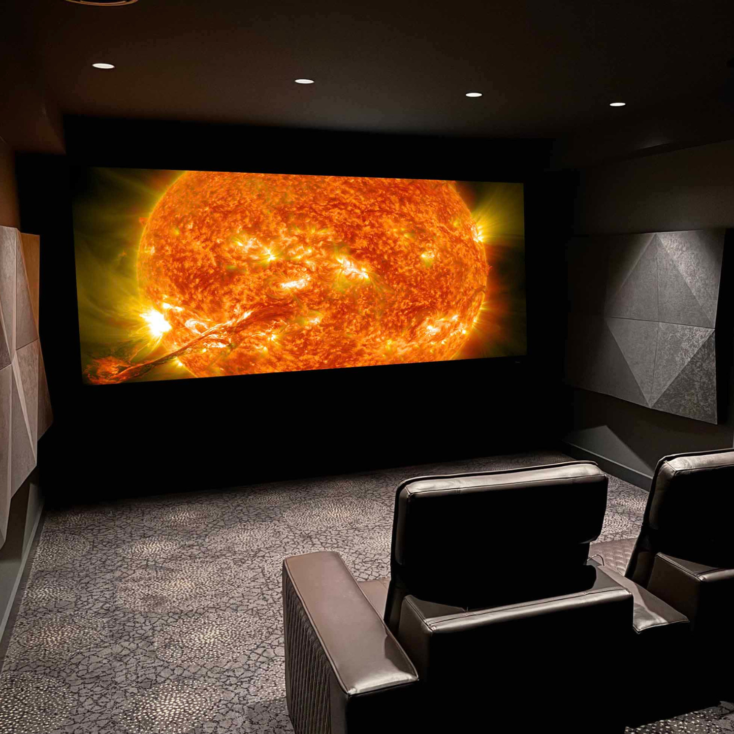 A home theater room with two leather recliner chairs facing a large wall-mounted screen displaying a close-up of the sun. The room has dark textured walls, patterned gray carpet, and recessed ceiling lights.