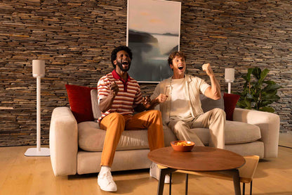Two people sitting on a couch in a modern living room, cheering or reacting to something off-camera.