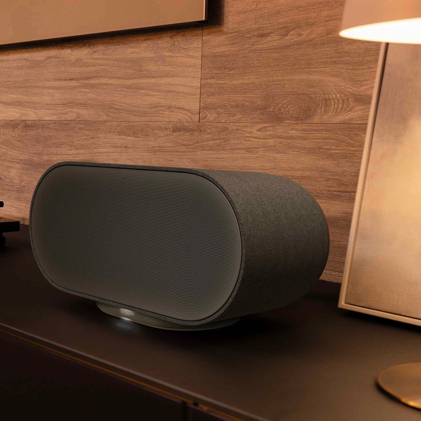 Black Denon Home 600 speaker on a wooden surface with a lamp in the background