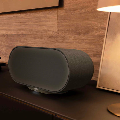 Black Denon Home 600 speaker on a wooden surface with a lamp in the background