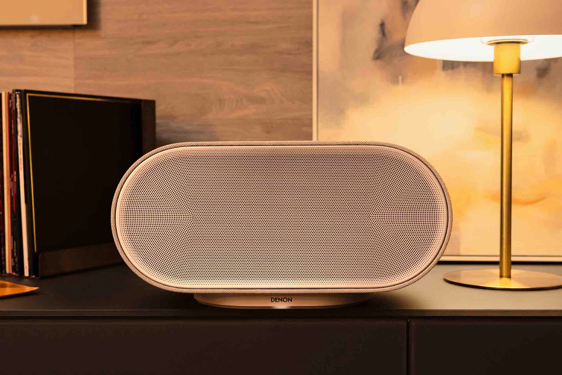 White Denon speaker on a wooden surface with a lamp and books in the background