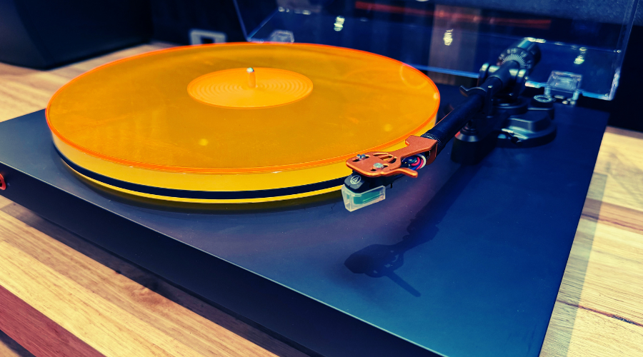 A close-up of a turntable with a translucent orange mat on a black plinth, photographed on a wooden surface.