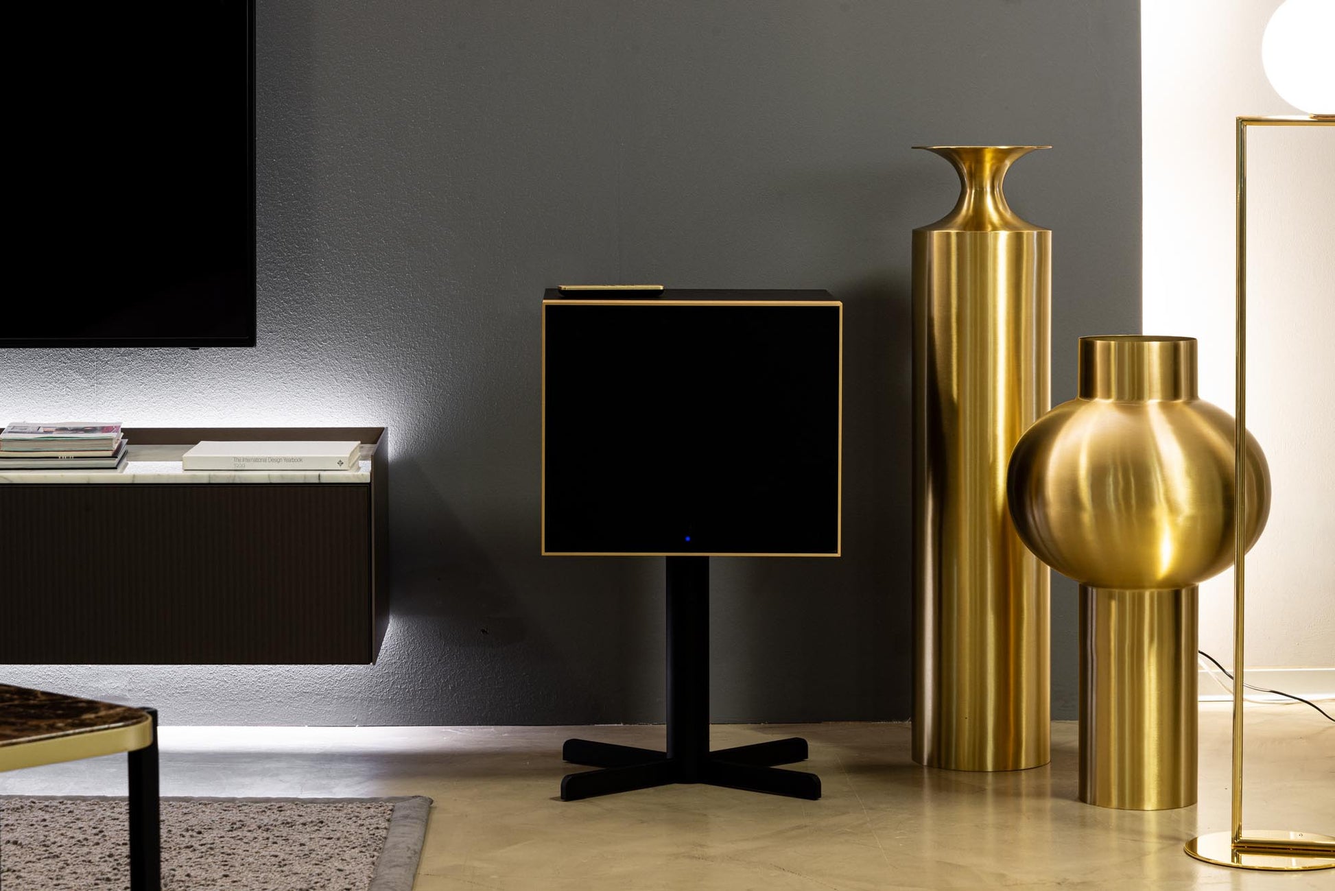 Modern interior with a black speaker on a stand, gold vases, and a gray wall.