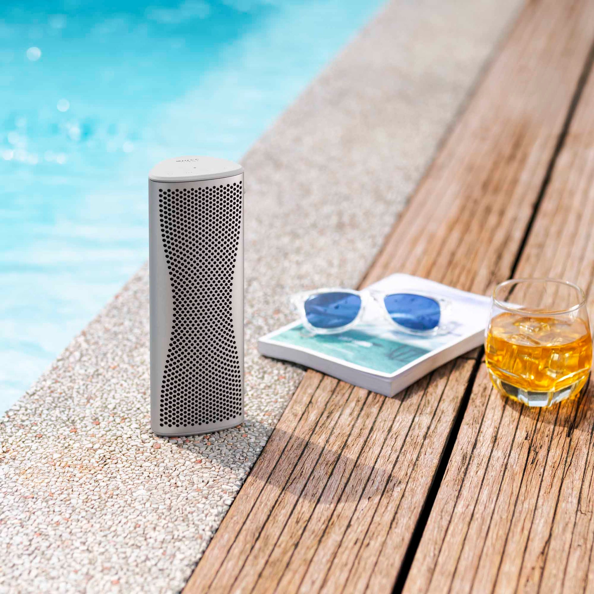 Portable speaker on a wooden deck by a poolside with sunglasses and a glass of orange juice.