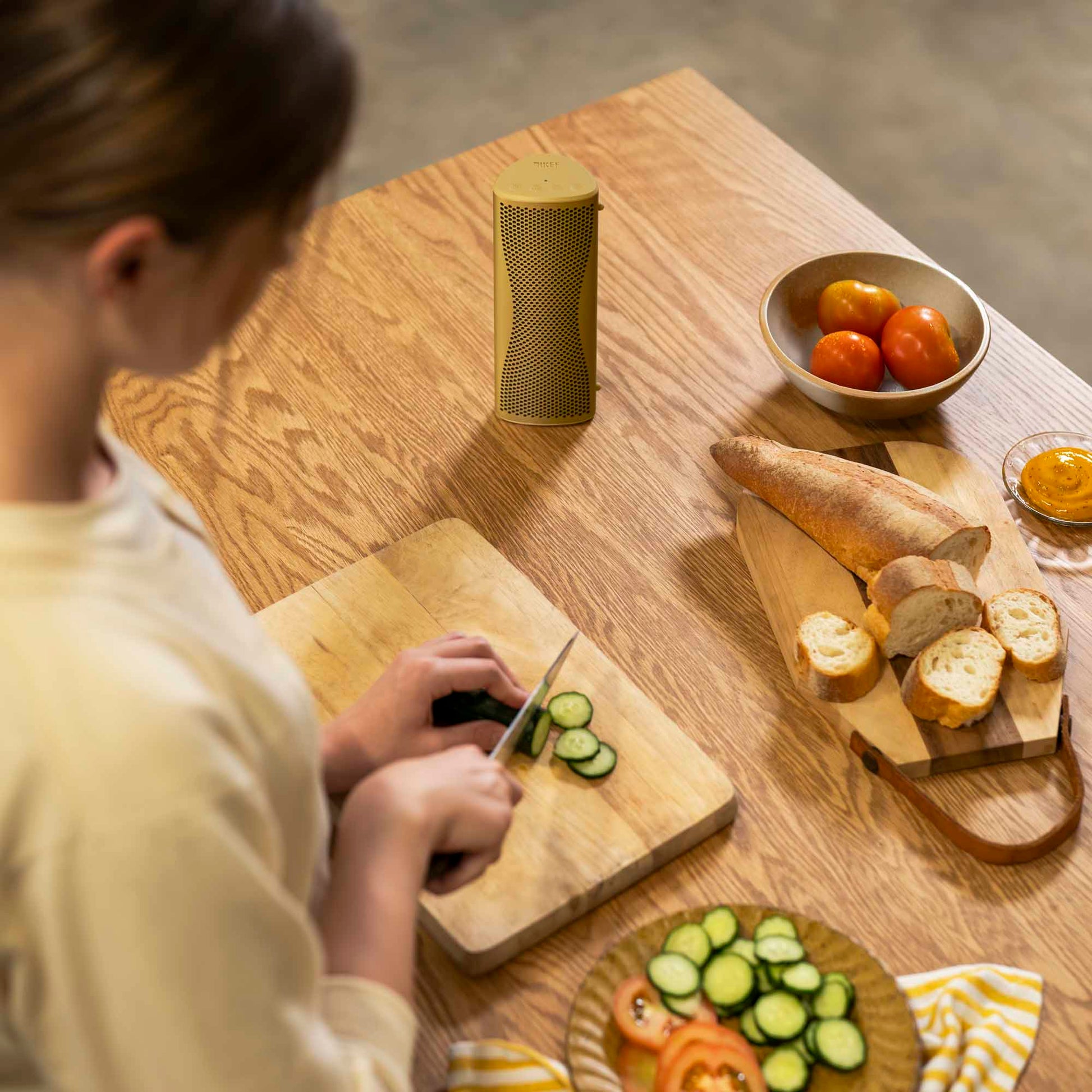Person slicing cucumbers on a wooden cutting board with bread and fruits on a table.