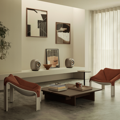 A modern living room scene with two sculptural orange-brown armchairs, a low dark wood coffee table, a floating shelf with decorative objects, and framed abstract wall art against a warm beige wall.