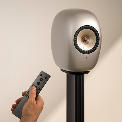 KEF LS LUXE Wireless Powered Speaker in dusk Titanium on a black stand, shown with a hand holding a remote control against a plain beige background.