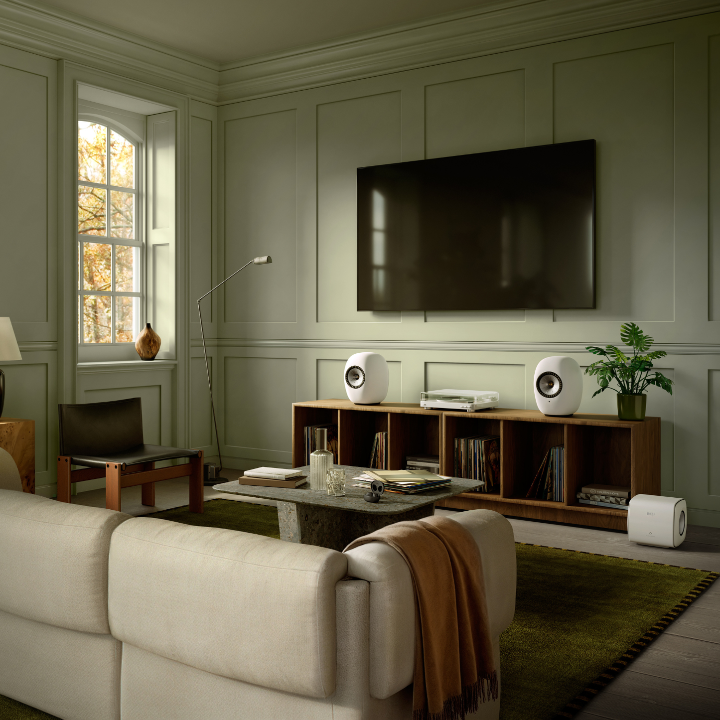 A modern living room with light sage green paneled walls, featuring a large black flat-screen TV mounted on the wall above a wooden media console. The room is styled with a cream-colored sofa in the foreground, white speakers on the console, a green plant, and a green area rug.
