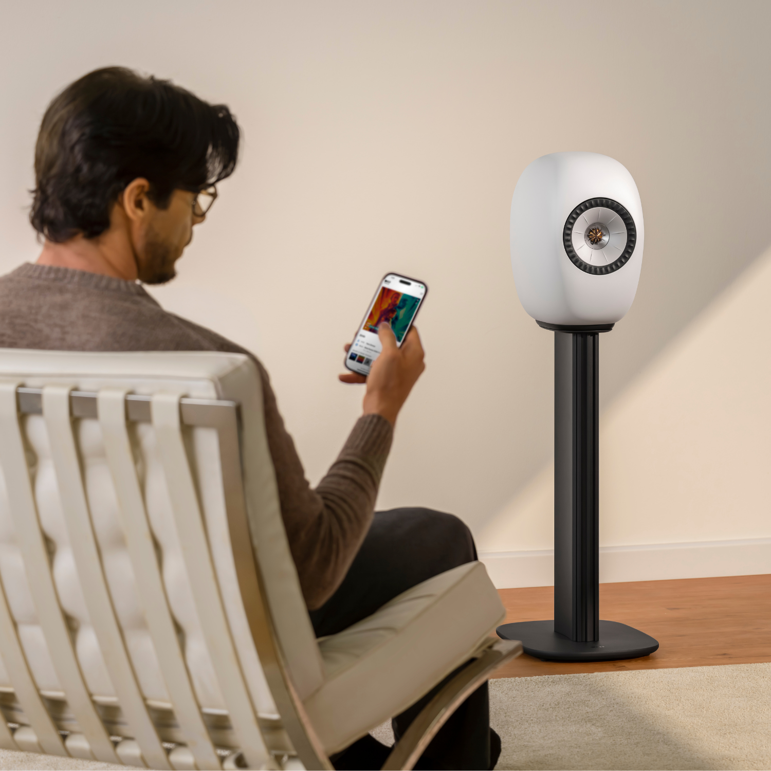 A man sits in a white lounge chair, holding a smartphone app screen, while a white wireless speaker stands on a stand in a minimalist room.