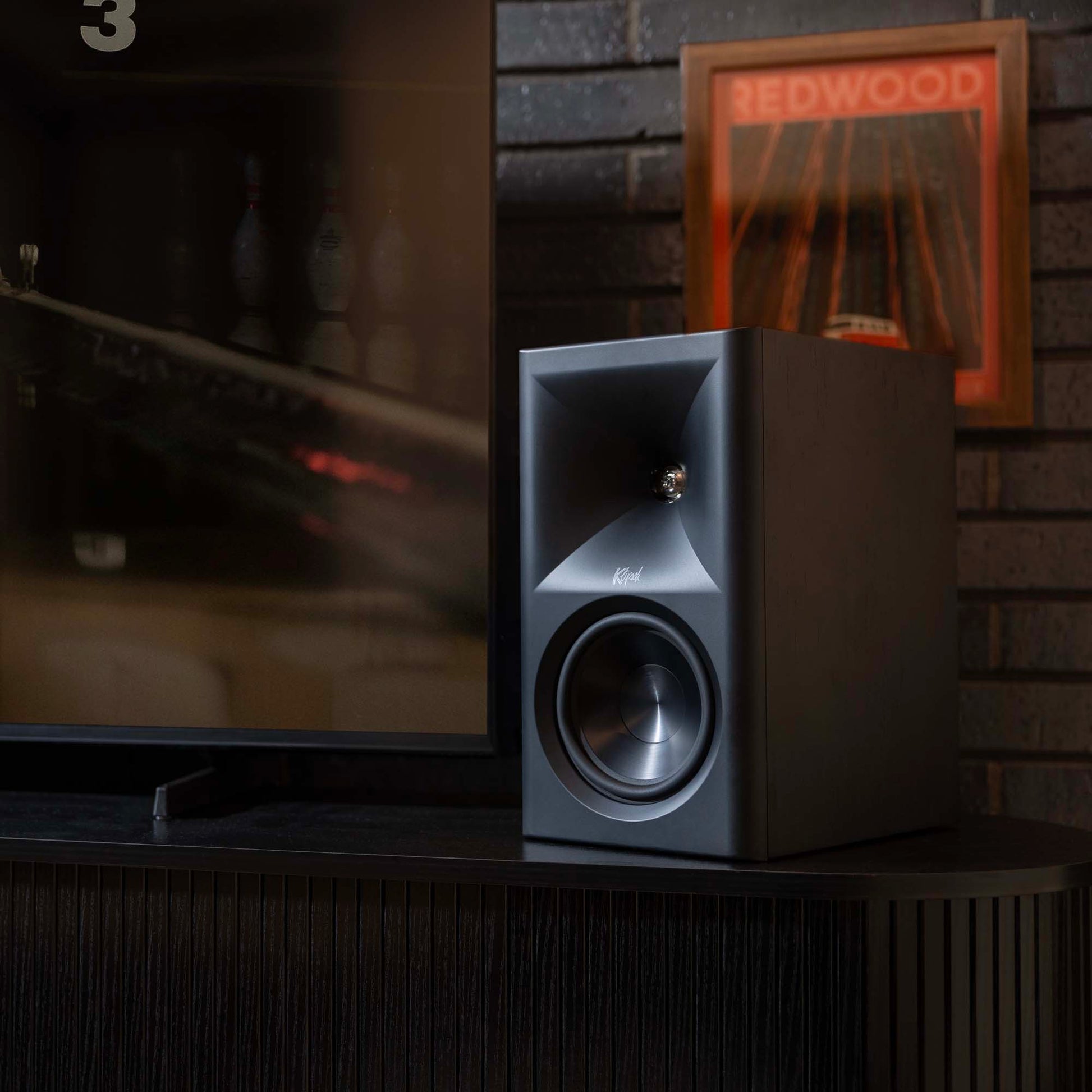 Speaker on a cabinet in a room with a television and framed picture in the background