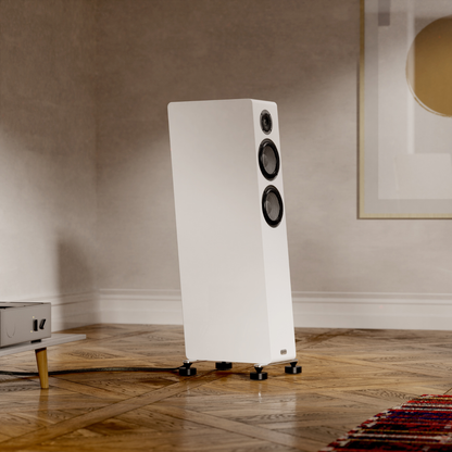 White floor-standing speaker in a room with wooden flooring and a neutral wall.