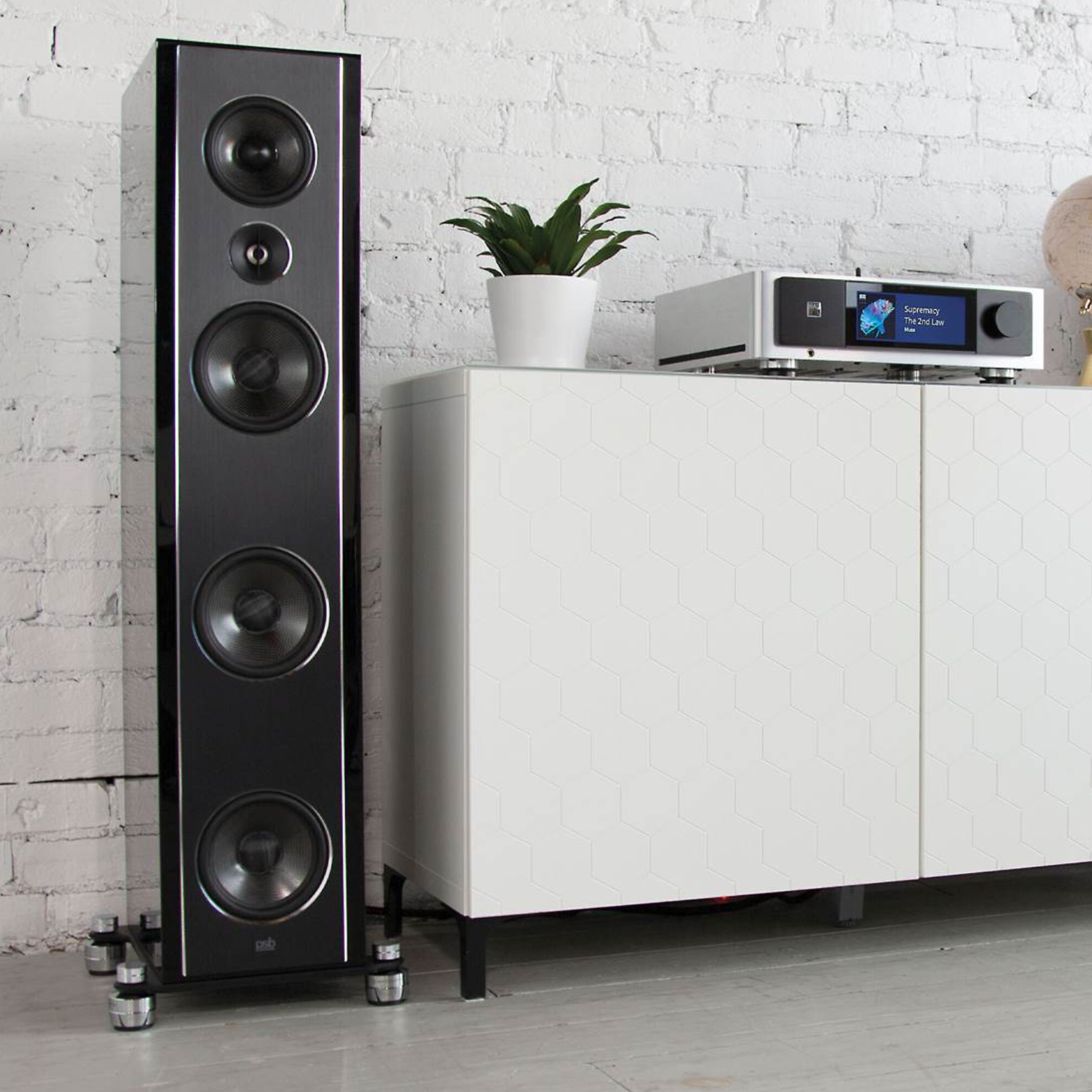 Black floor-standing speaker next to a white cabinet with electronic equipment on a light gray floor against a white brick wall.