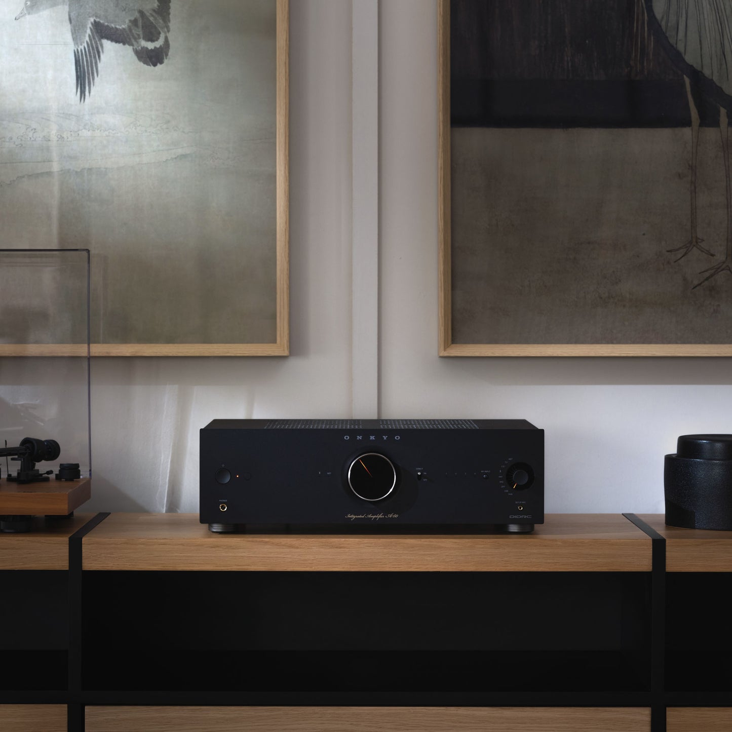 Black audio amplifier on a wooden surface with framed artwork in the background
