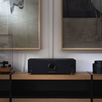 Black audio amplifier on a wooden surface with framed artwork in the background