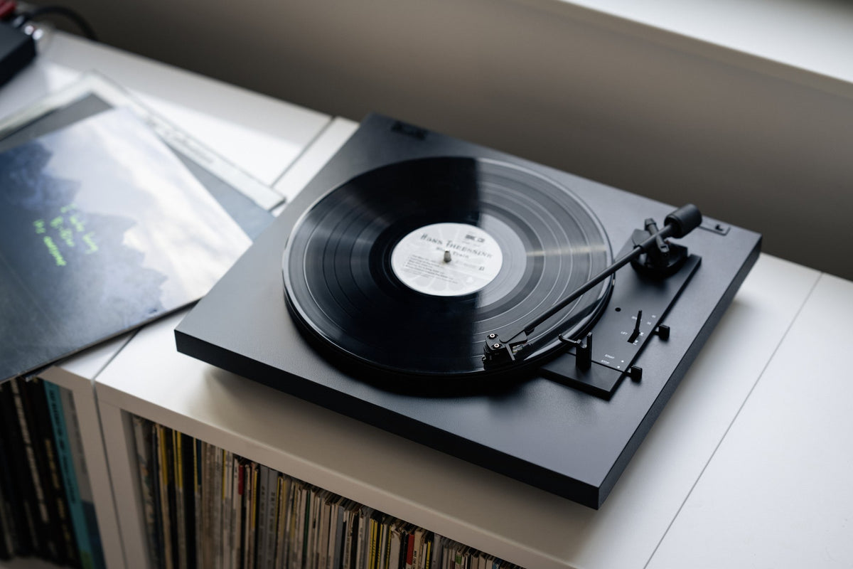 A black turntable with a vinyl record playing, set on a white shelf with a record rack and album sleeves in the background.