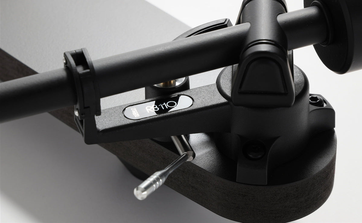 Close-up of a black turntable tonearm with a visible stylus and a small oval badge reading 'RB110' on a light gray background.